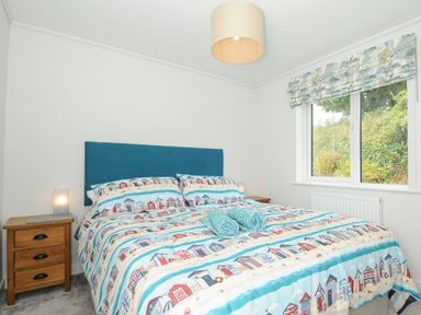 A bedroom with a bed with blue headboard and beach themed bedding two wooden nightstands a window with patterned blinds and a ceiling light at Midway Lodge in Cawsand near Kingsand