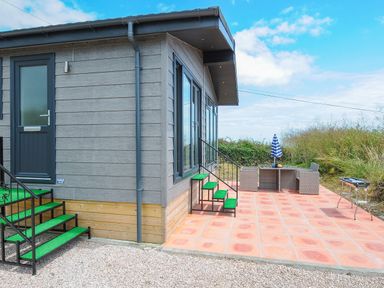 An outdoor patio area with tiled floor a table and chairs with umbrella and a barbecue next to a small building with green steps at Midway Lodge in Cawsand near Kingsand