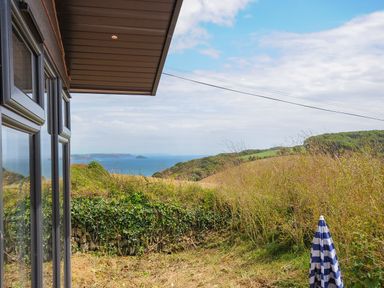 A view of grassy hills and the sea seen from a garden with a striped umbrella and house windows at Midway Lodge in Cawsand near Kingsand