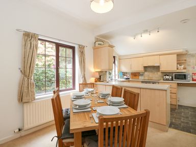 A dining area with wooden table and six chairs set with dishes beside a kitchen with light wood cabinets at Brantfell Lodge in Bowness-On-Windermere