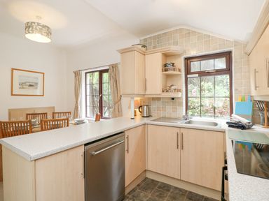 A kitchen with wooden cabinets stainless steel dishwasher two windows above sink and dining table with chairs at Brantfell Lodge in Bowness-On-Windermere