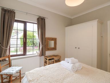 A bedroom with a bed covered in textured bedding and rolled white towels a wooden chair with a cushion a window with curtains a white wardrobe and a small shelf with baskets at Brantfell Lodge in Bowness-On-Windermere