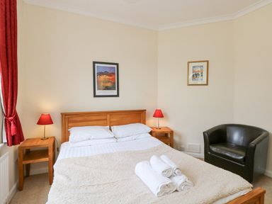 A bedroom with a double bed with white linens and towels on top two wooden bedside tables with red lamps a black armchair and framed pictures on the wall at Brantfell Lodge in Bowness-On-Windermere