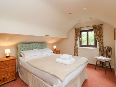 A bedroom with a bed with rolled towels bedside tables with lamps a window with floral curtains and a wooden chair with cushion at Brantfell Lodge in Bowness-On-Windermere