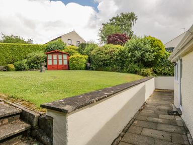 A garden area with a red shed surrounded by bushes and trees with a paved walkway and steps at Brantfell Lodge in Bowness-On-Windermere