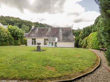 A lawn with a stone planter and a gravel path with a bench around a house surrounded by trees at Brantfell Lodge in Bowness-On-Windermere
