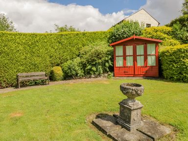 A garden with a stone planter on a pedestal a wooden bench and a red garden shed at Brantfell Lodge in Bowness-On-Windermere