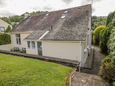 A side view of a two-story house with a tiled roof and a paved pathway surrounded by bushes at Brantfell Lodge in Bowness-On-Windermere