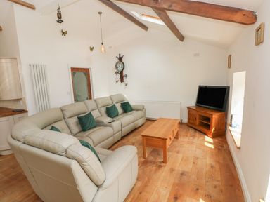 A living room with a beige leather sectional sofa and green pillows a wooden coffee table a wooden TV stand with a television exposed wooden ceiling beams and a clock on the wall at Greenheart Cottage in Pontrhydyfen