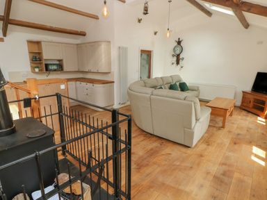 A living room with a beige sectional sofa green pillows a wooden coffee table a wood stove a black metal guard a TV on a wooden stand and a kitchen with wooden countertops at Greenheart Cottage in Pontrhydyfen