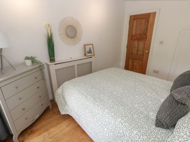 A bedroom with a bed covered in patterned bedding a dresser with a lamp and plant and a wooden door at Greenheart Cottage in Pontrhydyfen