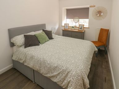 A bedroom with a double bed with patterned bedding a gray dresser with framed pictures a chair and a round mirror at Greenheart Cottage in Pontrhydyfen