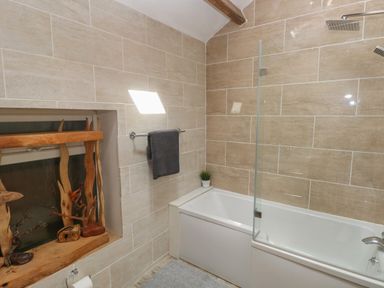 A bathroom with a bathtub and shower area featuring beige tiled walls and a wooden decoration near the window at Greenheart Cottage in Pontrhydyfen