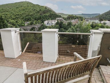 A balcony with a wooden bench overlooking a village and green hills at Greenheart Cottage in Pontrhydyfen