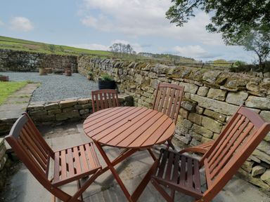 An outdoor seating area with a round wooden table and four chairs near a stone wall at The Hollow in West Woodburn