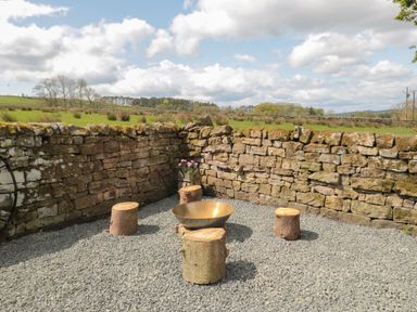 An outdoor seating area with tree stump stools and a fire bowl surrounded by stone walls at The Hollow West Woodburn