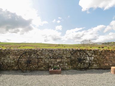 A stone wall with two metal wagon wheels and a rectangular planter with flowers in a gravel area at The Hollow in West Woodburn