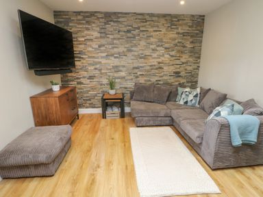 A living room with a gray sectional sofa, a wooden cabinet with a plant, a wall-mounted television, and a small table with a plant at Fir Tree Cottage in Llanrug