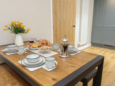 A dining table set with plates mugs a French press and baked goods in a kitchen at Fir Tree Cottage in Llanrug