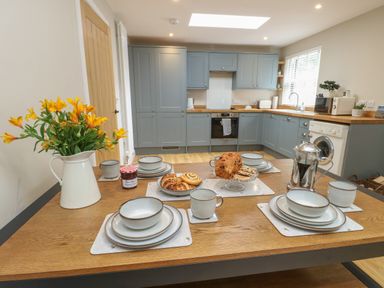 A kitchen with blue cabinets and a wooden dining table set with dishes pastries and a vase of yellow flowers at Fir Tree Cottage in Llanrug
