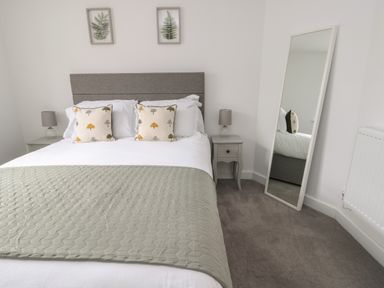 A bedroom with a bed with two pillows and a gray quilt a nightstand with a lamp a standing mirror and two framed botanical prints on the wall at Fir Tree Cottage in Llanrug