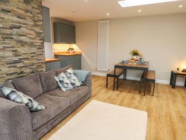 A living area with a grey sofa and patterned pillows a wooden dining table with benches and kitchen cabinets at Bay Tree Cottage in Llanrug