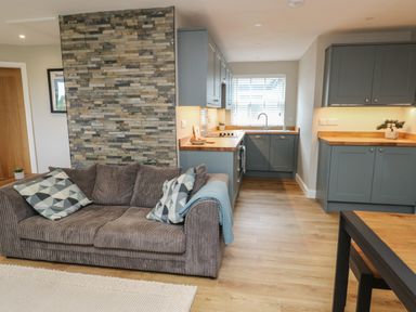 A living room with a brown sofa and cushions next to a kitchen with blue cabinets and wooden countertops at Bay Tree Cottage in Llanrug