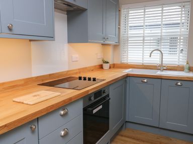 A kitchen with blue cabinets wooden countertops an electric stovetop oven a sink under a window and a small plant on the counter at Bay Tree Cottage in Llanrug