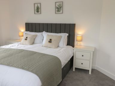 A bedroom with a grey upholstered bed headboard two bee design cushions a green quilt and white bedside tables with lamps at Bay Tree Cottage in Llanrug