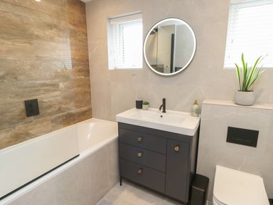 A bathroom with a bathtub with wooden wall tiles a black faucet and cabinet a round mirror and two windows at Bay Tree Cottage in Llanrug