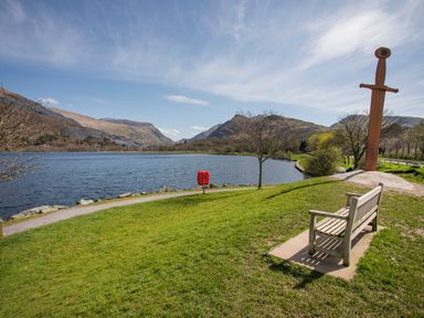A lake with a wooden bench and a large sword sculpture on grass with mountains in the background