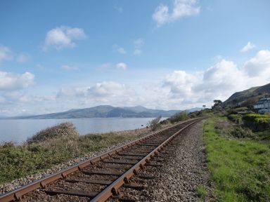 Railway tracks running alongside a body of water with hills in the background at Tawelfan in Llwyngwril