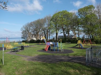 A children's playground with swings slides and climbing frames near trees and houses at Tawelfan in Llwyngwril