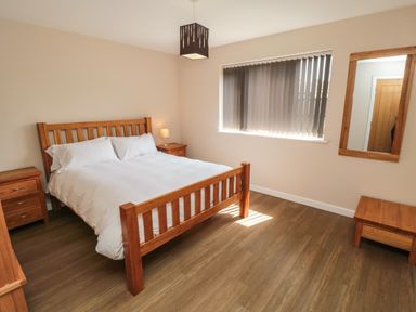 A bedroom with a wooden bed frame white bedding two wooden nightstands a window with vertical blinds a wall mirror and a wooden floor at 31A The Green in Scalby