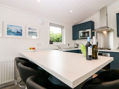 A kitchen with a white countertop bar stools bottles fruit bowl window and blue cabinets at Eden Fields Lodge in Cupar Fife