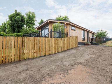 A wooden cabin with black railings and a wooden fence on a dirt ground at Eden Fields Lodge in Cupar Fife
