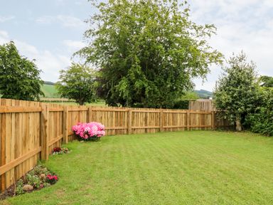 A fenced backyard with green grass pink flowers and trees at Eden Fields Lodge in Cupar Fife