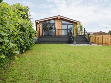 A wooden cabin with large windows and a black deck overlooking a grassy yard with a hedge on one side at Eden Fields Lodge in Cupar Fife