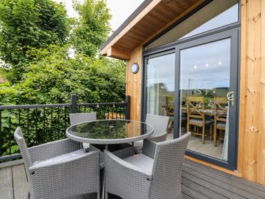 An outdoor wooden deck with a round glass table and four wicker chairs next to sliding glass doors at Eden Fields Lodge in Cupar Fife