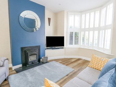 A living room with a blue accent wall fireplace a television on a corner shelf bay windows with white shutters and a gray sofa with yellow and blue cushions at Leos Place in Weymouth