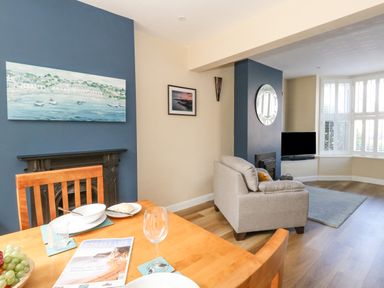 A dining and living area with a wooden table set for a meal a beige armchair a television and shuttered windows at Leos Place in Weymouth