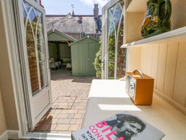 A table with a book and radio next to open glass doors leading to a paved yard with green garden sheds at Leos Place in Weymouth