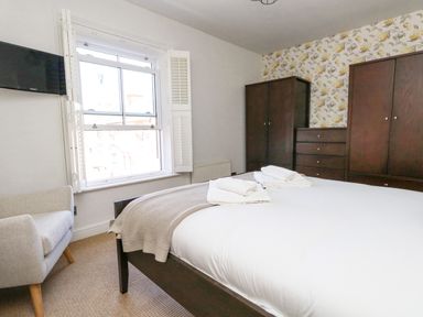 A bedroom with a bed towels on the bed a window a grey armchair dark wooden wardrobes and floral wallpaper at Leos Place in Weymouth