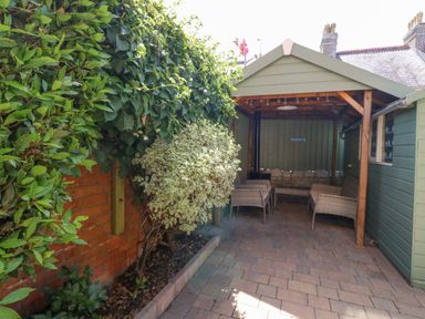 An outdoor covered seating area with wicker chairs and a small table next to green plants along a brick wall at Leos Place in Weymouth