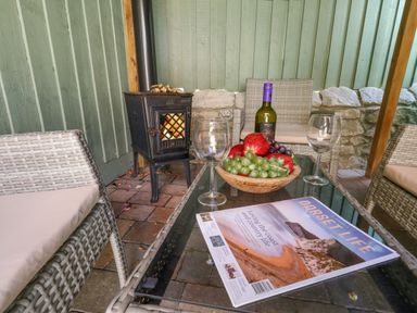 An outdoor seating area with wicker chairs a glass table a bowl of fruit a bottle of wine a magazine and a wood stove at Leos Place in Weymouth