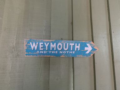 A blue wooden sign with white text pointing right on a wooden wall at Leos Place in Weymouth