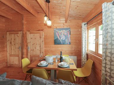 A dining area with a wooden table set for four yellow chairs two wooden doors and a window with patterned curtains at Pigsfoot Lodge in Ash Thomas near Halberton Devon