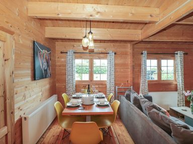 A dining area with a wooden table set for six and yellow chairs next to a gray sofa in a wooden cabin at Pigsfoot Lodge in Ash Thomas near Halberton Devon