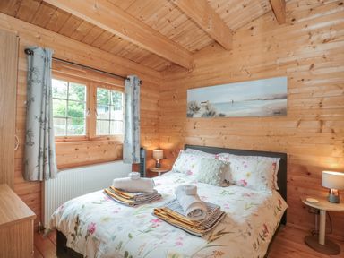 A bedroom with a floral bedspread and rolled towels on the bed wooden walls and ceiling with a window and curtains at Pigsfoot Lodge in Ash Thomas near Halberton Devon