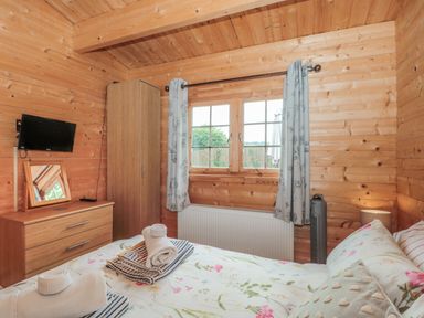 A bedroom with wooden walls a bed with floral bedding a chest of drawers a wardrobe a TV a window with curtains and a heater at Pigsfoot Lodge in Ash Thomas near Halberton Devon